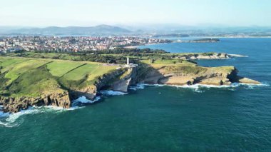 Arka planda yeşillik ve şehir manzarası olan kayalık bir sahildeki deniz fenerinin çarpıcı hava manzarası..