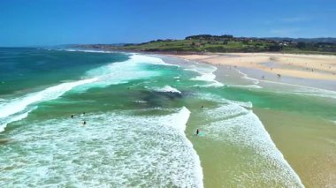 Hava manzarasında parlak ve güneşli bir günde turkuaz sularda sörf yapan sörfçülerin yer aldığı pitoresk bir plaj sergileniyor.