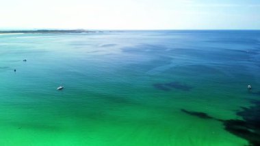 Açık mavi gökyüzünün altında sakin ve sakin bir turkuaz okyanusu gösteren nefes kesici bir hava görüntüsü.