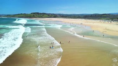 Güzel, canlı, çarpıcı dalgalar, güneş banyosu ve yemyeşil tepeler sergilenen bir plaj sahnesi.