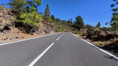 Dolambaçlı bir yol berrak mavi gökyüzünün altında parlak yeşilliklerle bezenmiş çarpıcı dağ manzarasının içinden geçiyor.