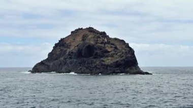 Dalgalar ve bulutlu gökyüzü ile çevrili, büyüleyici kayalık bir ada okyanustan yükseliyor..