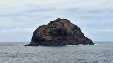 Sersemletici kayalık bir ada, sakin okyanus sularında, hepsi güzel, bulutlu bir havanın altında yer alır.