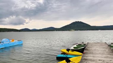 Güzel bir göl manzarası, canlı kanolar, güçlü bir rıhtım ve bulutlu bir gökyüzünün altında görkemli dağlar.