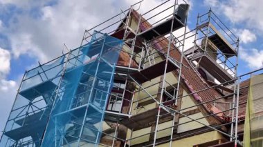 Şu anda açık mavi bir gökyüzünün altında onarım aşamasındaki büyük bir binanın iskelesinin detaylı bir görüntüsü.