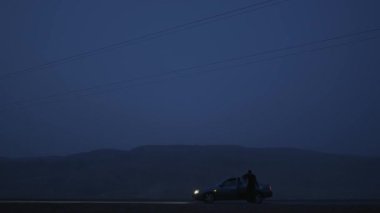 Gece bir dağ yolunda bir araba duruyor ve bir adam arabadan iniyor.