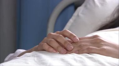 Hastane yatağında uzanmış kadının elini tutan bir adam, yakın plan el çekimleri.
