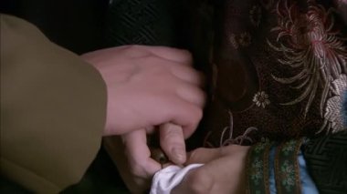 Adam annesinin elini tutuyor, anne de diğer elinde beyaz bir mendil tutuyor ve adamın eline koyuyor, yakın plan.