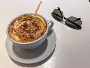 Üzerine kakao serpiştirilmiş köpüklü bir kapuçino ve beyaz bir masanın üzerinde şık bir güneş gözlüğü. Rahatlamış bir atmosfer izlenimi uyandırıyor..