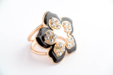 Parlak siyah taç yaprakları ve metalik bir zemine sabitlenmiş parlak parlak taşlardan oluşan çiçek şeklinde bir yüzüğün yakın çekimi. Yumuşak ışıklandırma ve ince detay ile beyaz bir arkaplanda fotoğraflandı.