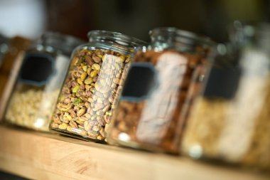 Cam Ceviz ve Tohum Kavanozları Ahşap bir rafta sergilenen şam fıstığı ve badem de dahil olmak üzere çeşitli fıstıklarla dolu bir sıra cam kavanoz. Bulanık arka plan sıcak ve davetkar bir atmosfer yaratıyor. Bu da bunu organik bir imaj haline getiriyor. 