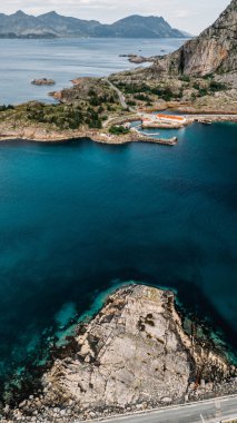 Norveç 'teki adalar ve uzak dağlarla engebeli kıyı şeridi boyunca uzanan arabalı dolambaçlı bir kıyı yolunun havadan görünüşü. Lofoten Adaları. Ağustos 2024