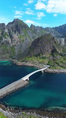 Norveç 'in Lofoten Adaları' nda dramatik dağlar ve yeşil tepeler tarafından çerçevelenmiş canlı bir mavi koy boyunca uzanan beyaz bir köprünün havadan görüntüsü..