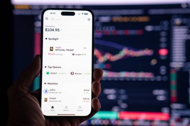 Elinde OFFICIAL TRUMP mim 'i gösteren bir telefon tutan adam, arka planda mim değerindeki dalgalanmayı gösteren bulanık bir monitörle. Budapeşte, Macaristan - 20 Ocak 2025