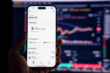 Elinde OFFICIAL TRUMP mim 'i gösteren bir telefon tutan adam, arka planda mim değerindeki dalgalanmayı gösteren bulanık bir monitörle. Budapeşte, Macaristan - 20 Ocak 2025