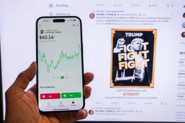 Man holding a phone displaying the official Trump meme logo in the Moonshot app, ready for purchase, with a blurred monitor in the background showing the meme's chart. Budapest, Hungary - jan 20, 2025 clipart