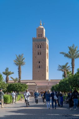 Koutoubia Camii - Marakeş, Fas, Afrika 'daki en büyük cami. Palmiyeli bahçelerin manzarası. Müslüman kıyafetleriyle yürüyen insanlar ve motosikletçiler..