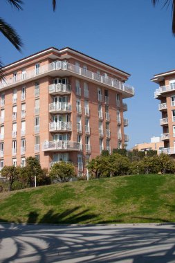 Sıcak tuğla cepheleri, beyaz korkuluklar ve birden fazla balkonu olan Akdeniz mimarisi tarzında modern bir apartman dairesi. Berrak mavi bir gökyüzü, yemyeşil çimenler ve ağaçlarla, sakin şehir atmosferine ekleniyor..