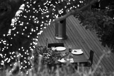 Close-up of a set table on a patio, with illuminated tree branches in monochrome, creating a warm ambiance. Outdoor dining, decorative lighting, and a romantic setting. clipart