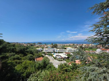 Yeşil ağaçlar ve deniz tarafından çerçevelenmiş canlı bir Yunan turizm kasabasına bakan manzaralı bir manzara.