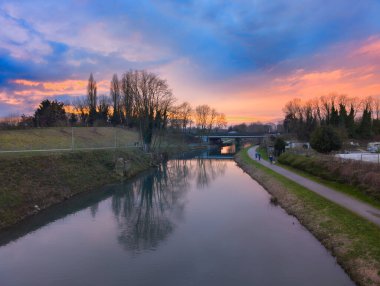 Gün batımında dingin bir kanal sahnesi. Suyu yansıtan renkli bir gökyüzü. Ağaçlarla çevrili ve yürüyen bir patika..