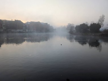 Suyu ve uzak binaları sisli bir sis kaplayan sakin bir nehir manzarası. Sakin su bulutlu gökyüzünü yansıtır ve sakin bir atmosfer yaratır. Ağaçlar ve binalar nehir kıyısında..
