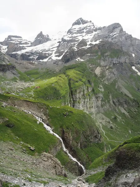 Şelale, arka planında karlı dağlar olan yeşil bir vadiye akar, İsviçre. Yaz boyunca İsviçre Alpleri