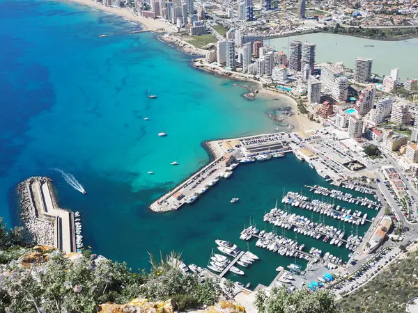 Beyaz yatlı Calpe limanı manzarası, Costa Blanca, Alicante, İspanya. Kumsal boyunca gökdelenler ve güneşli bir günde mavi Balear Denizi. Tekne sakin mavi sularda yol alır, arkasında beyaz bir iz bırakır..