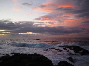 Atlantik Okyanusu sahilinde güzel bir günbatımı: pembe yumuşak ışık, mavi dalgalar, kayalık kara kıyı ve ufukta bulutlar. Mesa del Mar, Tenerife, Kanarya Adaları, İspanya.