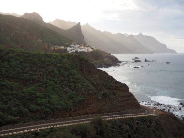Atlantik kıyısındaki Kuzey Tenerife 'nin yeşil volkanik kıyısındaki Roque de las Animas üçgen kayasının altındaki beyaz köy. İspanya 'nın Kanarya Adaları' ndaki uçurumların tepelerinden gün batımı ışığı yükseliyor..