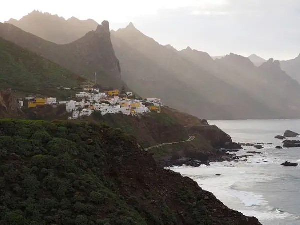 Atlantik kıyısındaki Kuzey Tenerife 'nin yeşil volkanik kıyısındaki Roque de las Animas üçgen kayasının altındaki beyaz köy. İspanya 'nın Kanarya Adaları' ndaki uçurumların tepelerinden gün batımı ışığı yükseliyor..