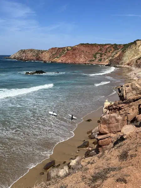 Atlantik Okyanusu 'ndaki sörfçüler Portekiz' in Algarve kıyısındaki kayalık bir sahilde dalgaları bekliyorlar. Turkuaz denizin, dalgaların ve açık mavi gökyüzünün altındaki kıyı kayalıklarının manzarası, Carrapateira