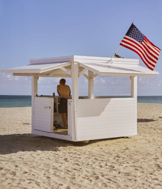Fotoğraf, parlak güneşin altında el değmemiş bir Miami plajında ayakta duran bir cankurtaran kulesini yakalıyor. Beyaz renklere boyanmış cankurtaran kulübesi, Amerikan bayrağıyla sakin, derin mavi bir denizin arka planına karşı odak noktasıdır.. 