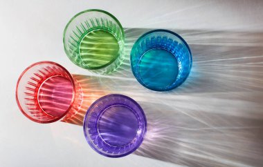 Görüntü, yukarıdan bakıldığında beyaz bir yüzeye yerleştirilmiş dört renkli içki bardağını gösteriyor. Gözlükler kırmızı, yeşil, mavi ve mordur. Her biri ışıktan dolayı karmaşık ve canlı gölgelerdir.. 