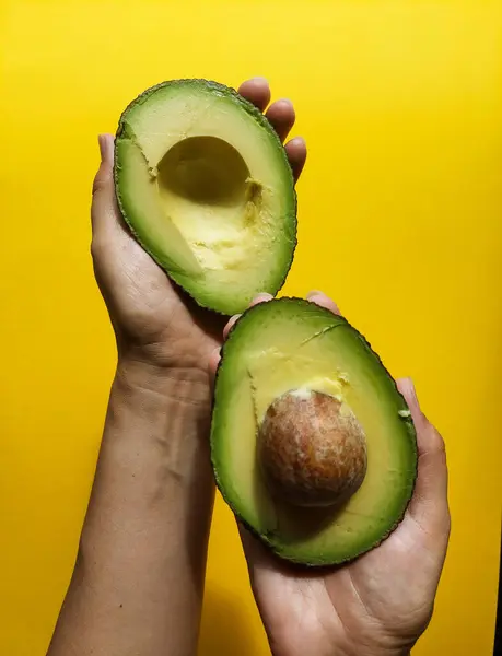 Sarı arka planda bir avokadonun iki yarısını tutan eller.