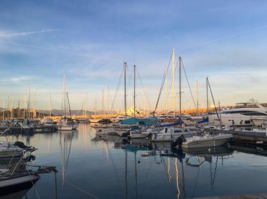Gün batımında marinanın güzel bir manzarası, sakin suları yansıtan çeşitli tekne ve yatlar. Mavi gökyüzü, gün batımının ılık tonlarıyla harmanlanır..