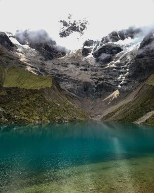 El değmemiş turkuaz bir gölün göz kamaştırıcı manzarası kar kaplı zirvelerin altında kısmen sis bulutlarıyla örtülmüş, doğanın dingin ve el değmemiş güzelliğini yakalıyor..