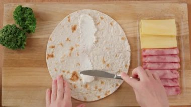 Tortilla ahşap bir tahtanın üzerinde yatar, kızların elleri tortillayı krem peynirle yayar ve jambonu serer.