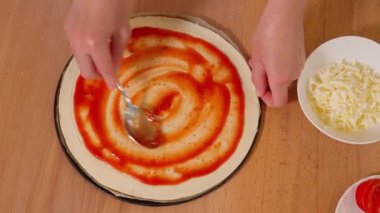 Tahta bir masada pizza pişirmek. Kızın elleri domates sosunu bir kaşıkla pizza hamurunun üzerine serp ve üzerine peynir serp. Pizza yiyecekler. Üst görünüm, yakın çekim