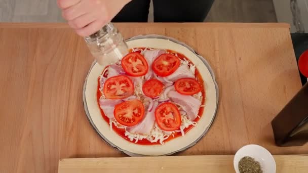 木のテーブルでピザを調理する 少女の手はミルを黒コショウに変え 芳香族のハーブを振りかけ ベーコンとトマトでピザにオリーブオイルを注ぐ トップビュー クローズアップ — ストック動画