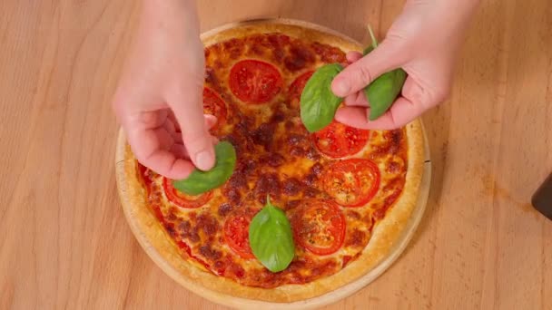 木のテーブルでピザを調理する 少女の手は完成したマルゲリータのピザにバジルを置き オリーブオイルを注ぎます トップビュー クローズアップ — ストック動画