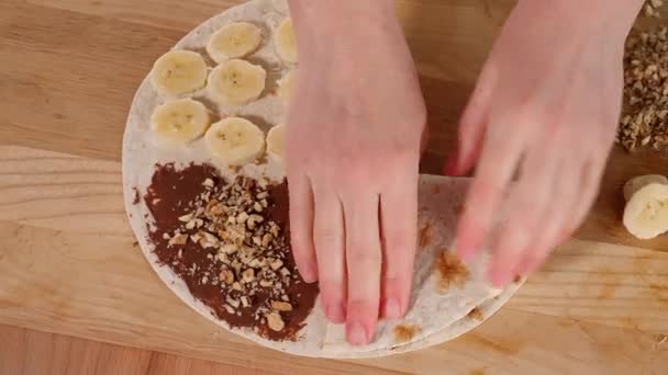 女孩用巧克力准备健康的素食早餐 用坚果 香蕉和木桌铺着比塔面包 圆圆的玉米饼被分成四个填充物 女孩们的手把玉米饼折叠成一个信封 — 图库视频影像