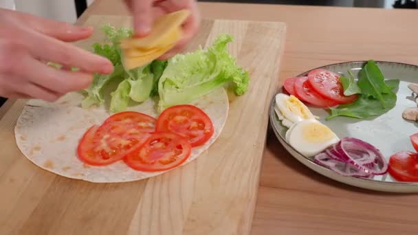 ピタパンと野菜 木のテーブルで健康的なビーガン朝食を準備する少女 トマトのスライスとチーズのスライスをサラダと丸トルティーヤに置き 封筒に折ります クローズアップ サイドビュー — ストック動画
