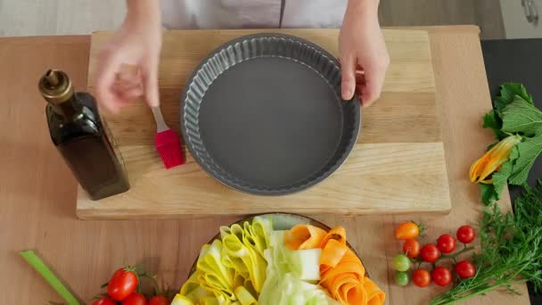 厨师把一个烘烤盘放在一张木制桌子上 用橄榄油刷着 背景中夹着西葫芦和西红柿 — 图库视频影像