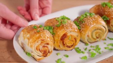 Pastırma ve peynirli taze pişmiş çörekler, susam tohumları ve yeşil soğanlar serpiştirilmiş, beyaz tabakta, yakın plan, ön manzara, makro. Kızın elleri bir tabak çöreği daire şeklinde çeviriyor..