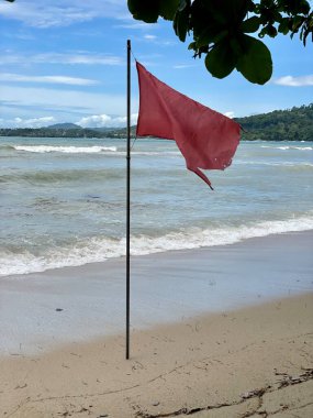 Deniz kıyısındaki kumların üzerinde kırmızı bir bayrak duruyor mavi gökyüzünün arka planında dağınık bulutlar ve tehlikeli dalgalarla dolu bir deniz..