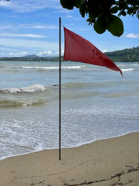 Deniz kıyısındaki kumların üzerinde kırmızı bir bayrak duruyor mavi gökyüzünün arka planında dağınık bulutlar ve tehlikeli dalgalarla dolu bir deniz..