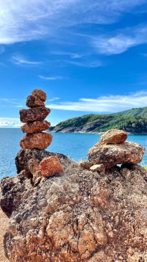 Deniz manzaralı, yeşil tepeler ve mavi gökyüzü olan kayalık bir sahilde dengeli taşlar. Meditasyon ve doğa elementleriyle sakin bir tropikal manzara..