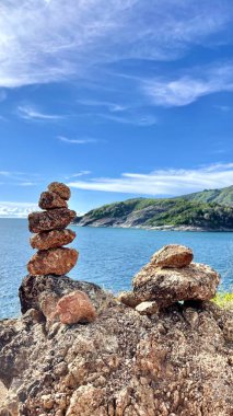 Deniz manzaralı, yeşil tepeler ve mavi gökyüzü olan kayalık bir sahilde dengeli taşlar. Meditasyon ve doğa elementleriyle sakin bir tropikal manzara..