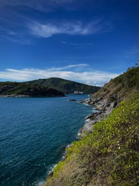 Deniz manzarası ve kayalık sahil. Arka planda mavi gökyüzünün altındaki yeşil ada. Konsept: Seyahat, doğa, tatil, tropikal cennet, eko-turizm, deniz özgürlüğü, bilgisayar duvar kağıdı.